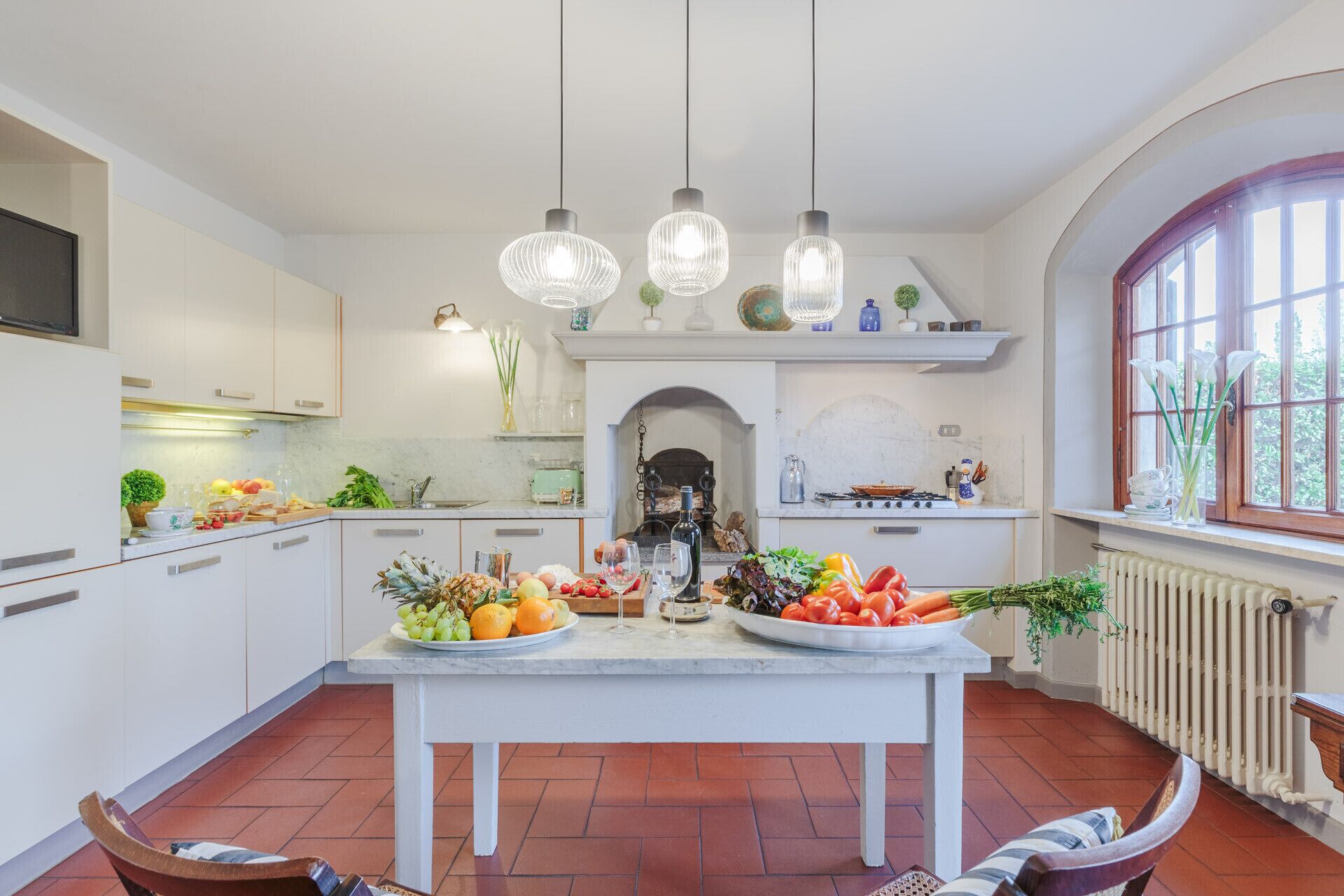 Une cuisine moderne avec des meubles blancs, un îlot central agrémenté de fruits et de boissons, des luminaires suspendus et une vue sur l’extérieur grâce à une fenêtre.