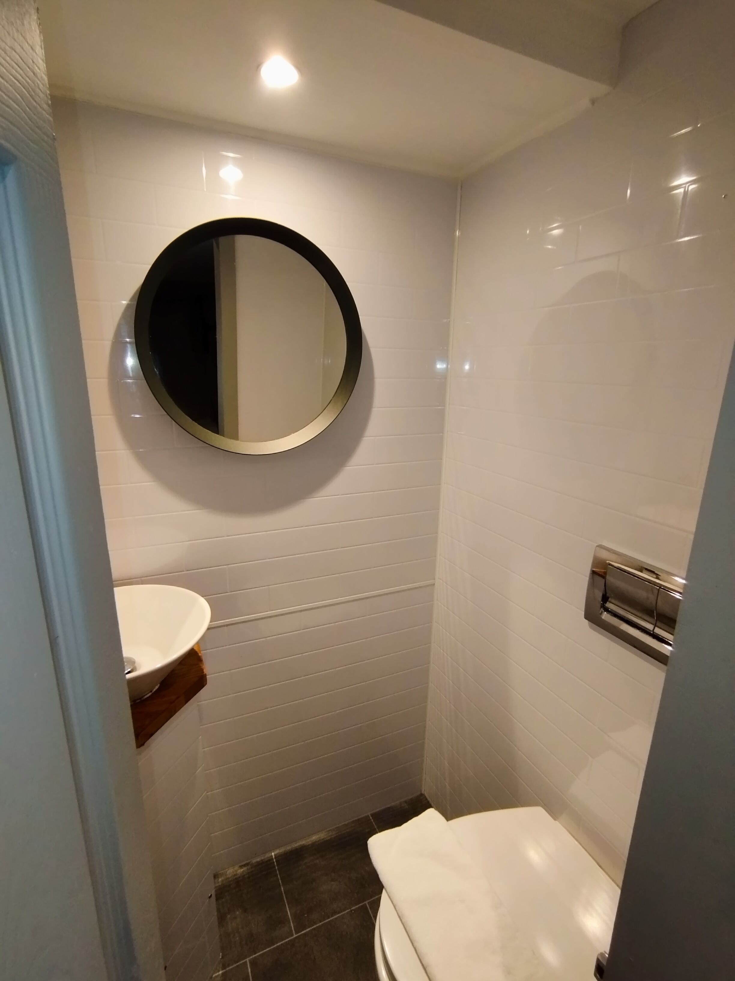 A small, modern bathroom with white tiled walls, a round mirror, a white sink, and a toilet.
