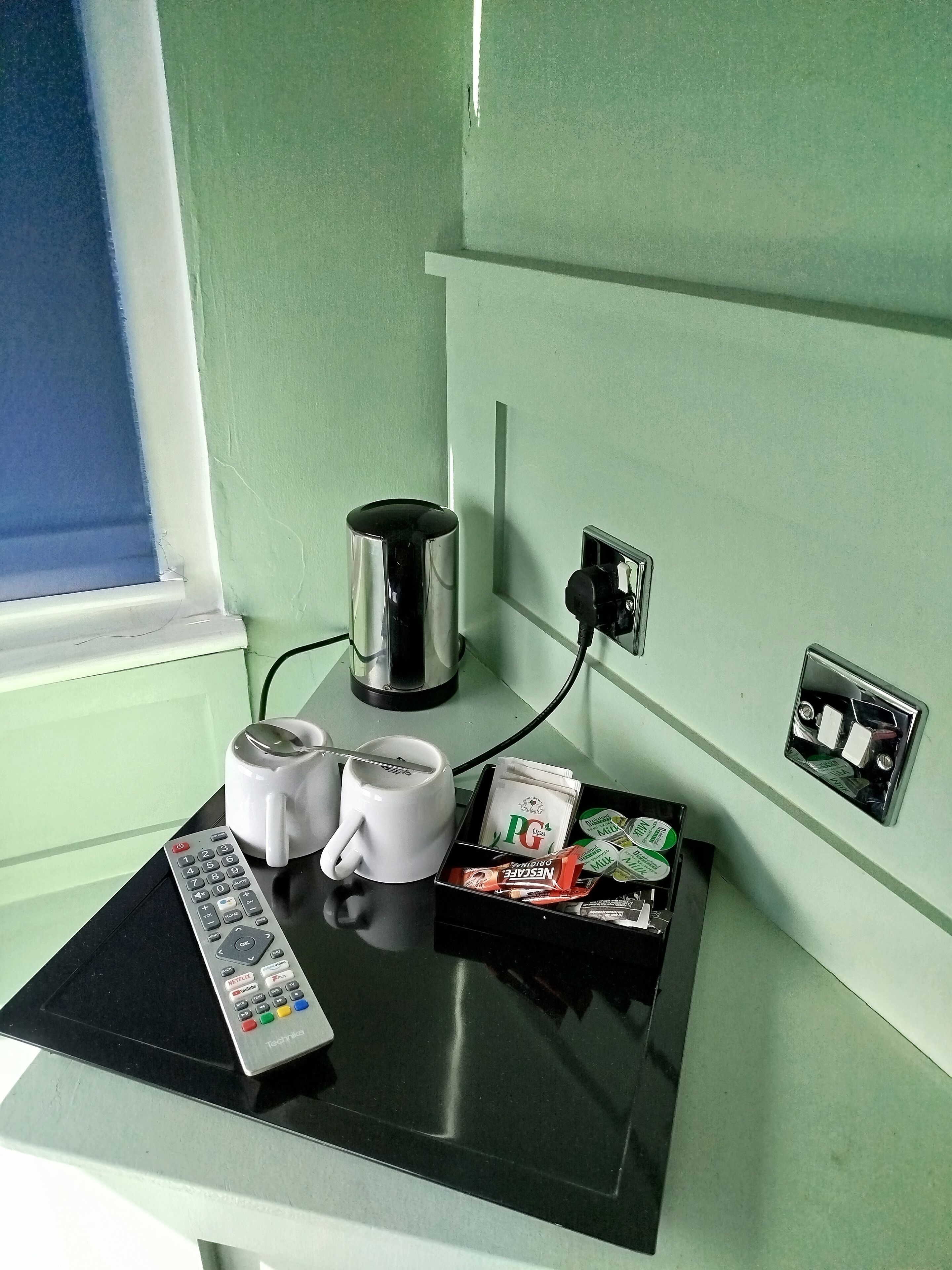 A small kitchenette with a coffee maker, two mugs, a remote control, and a tray of snacks.