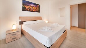 Quarto de hotel moderno com cama de madeira, roupa de cama branca, mesa de cabeceira com abajur e um quadro com vista para a cidade na parede.