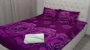 A bed with a purple floral bedspread and matching pillows, a white tufted headboard, and a folded towel on the bed. - 2 Bed-aircon-free Parking-city Centre (Chiajna)
