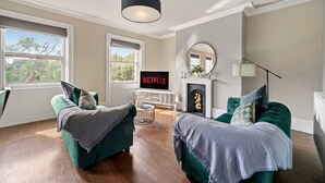 Una sala de estar moderna con dos sofás verdes, una chimenea, un televisor mostrando 'NETFLIX' y un ventanal grande.