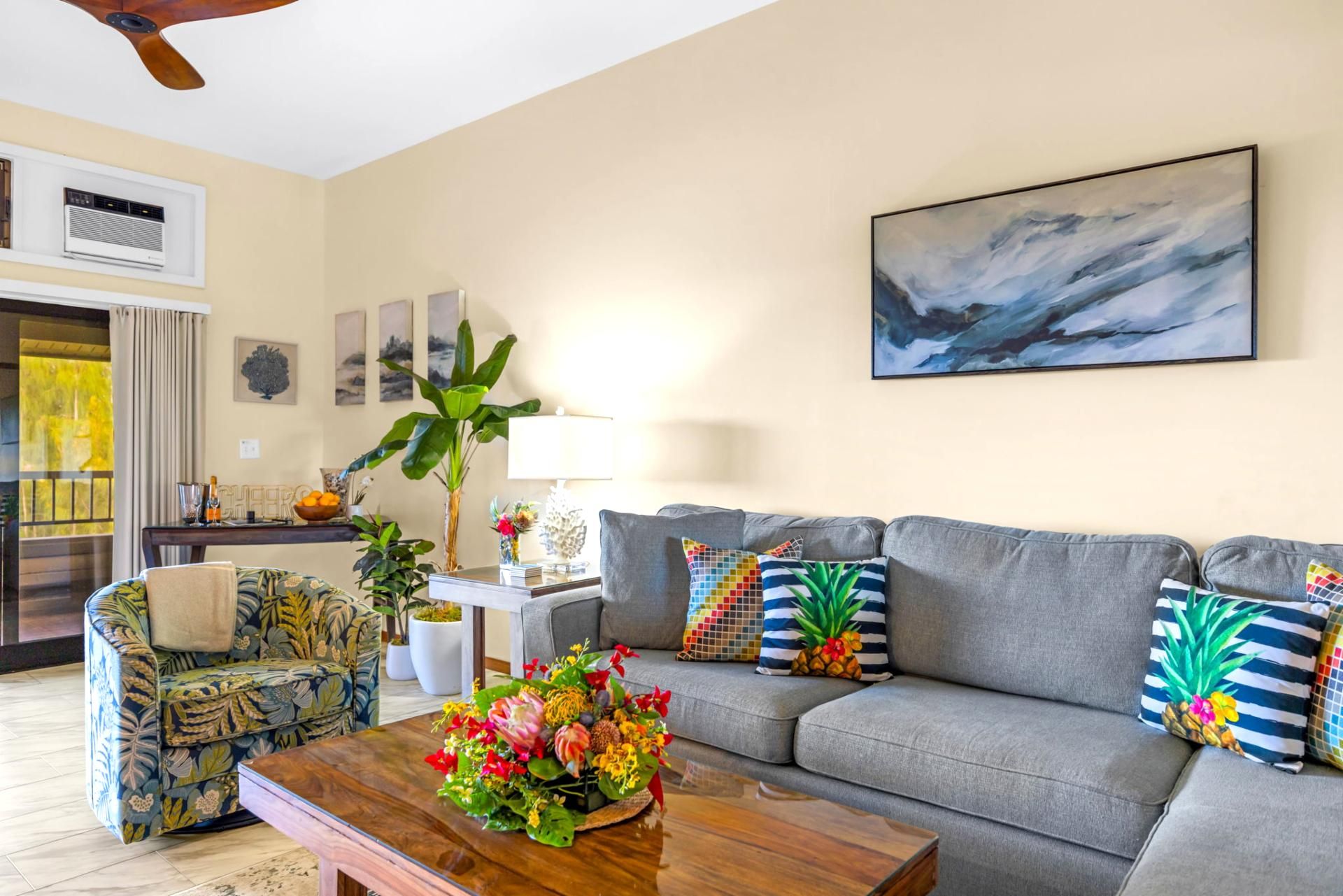 A living room with a grey sofa, floral armchair, and a coffee table with a floral arrangement. There is a painting on the wall and a window with curtains.