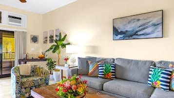 A living room with a grey sofa, floral armchair, and a coffee table with a floral arrangement. There is a painting on the wall and a window with curtains.