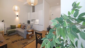 A modern living room with a grey sofa, a coffee table, and a dining area with a table and chairs. There is a kitchen area with white cabinets and a hanging light fixture. - FLH Gaia Homey Winery Flat (Vila Nova de Gaia)