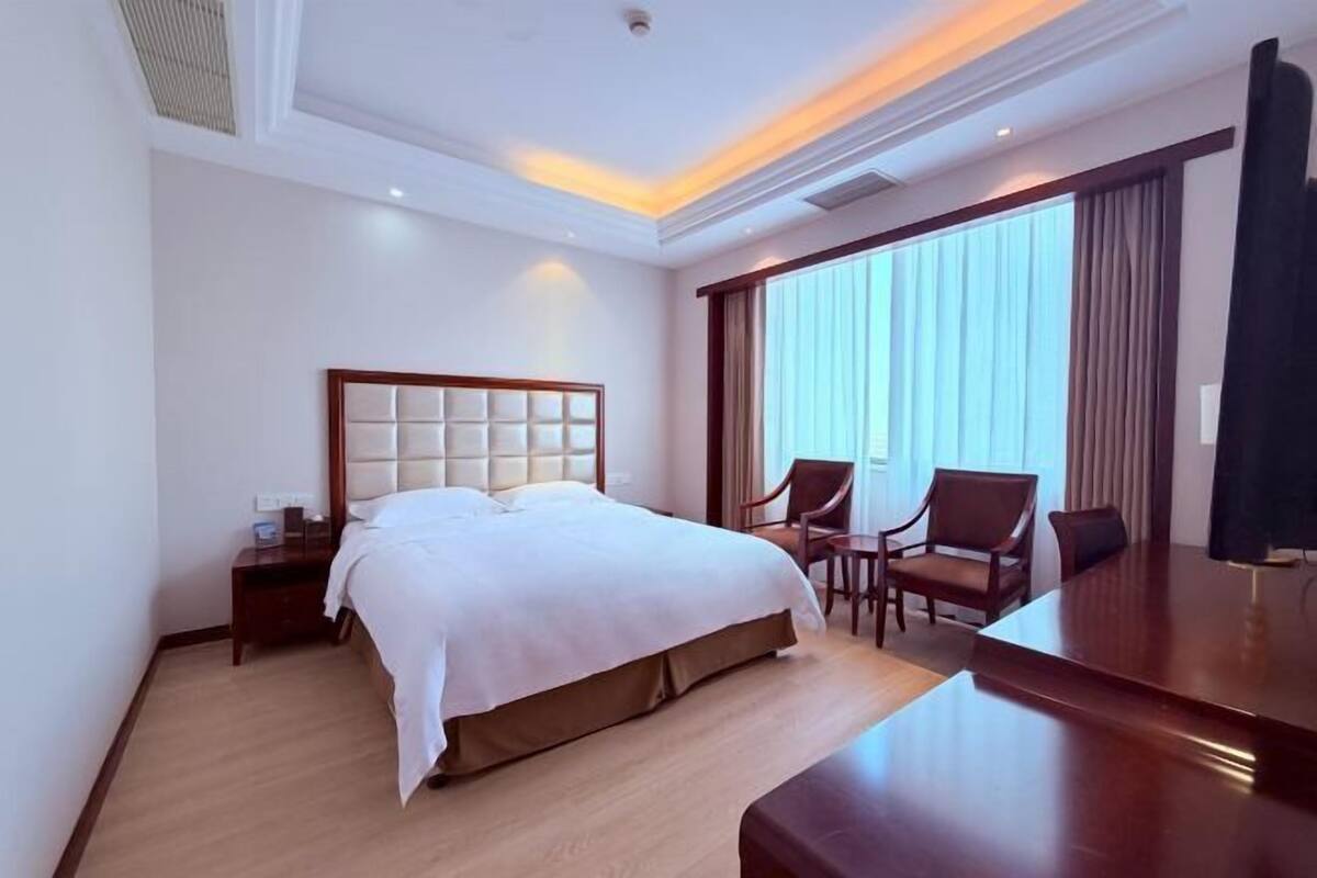 Habitación de hotel con una cama grande, un escritorio, dos sillas y un ventanal con cortinas.