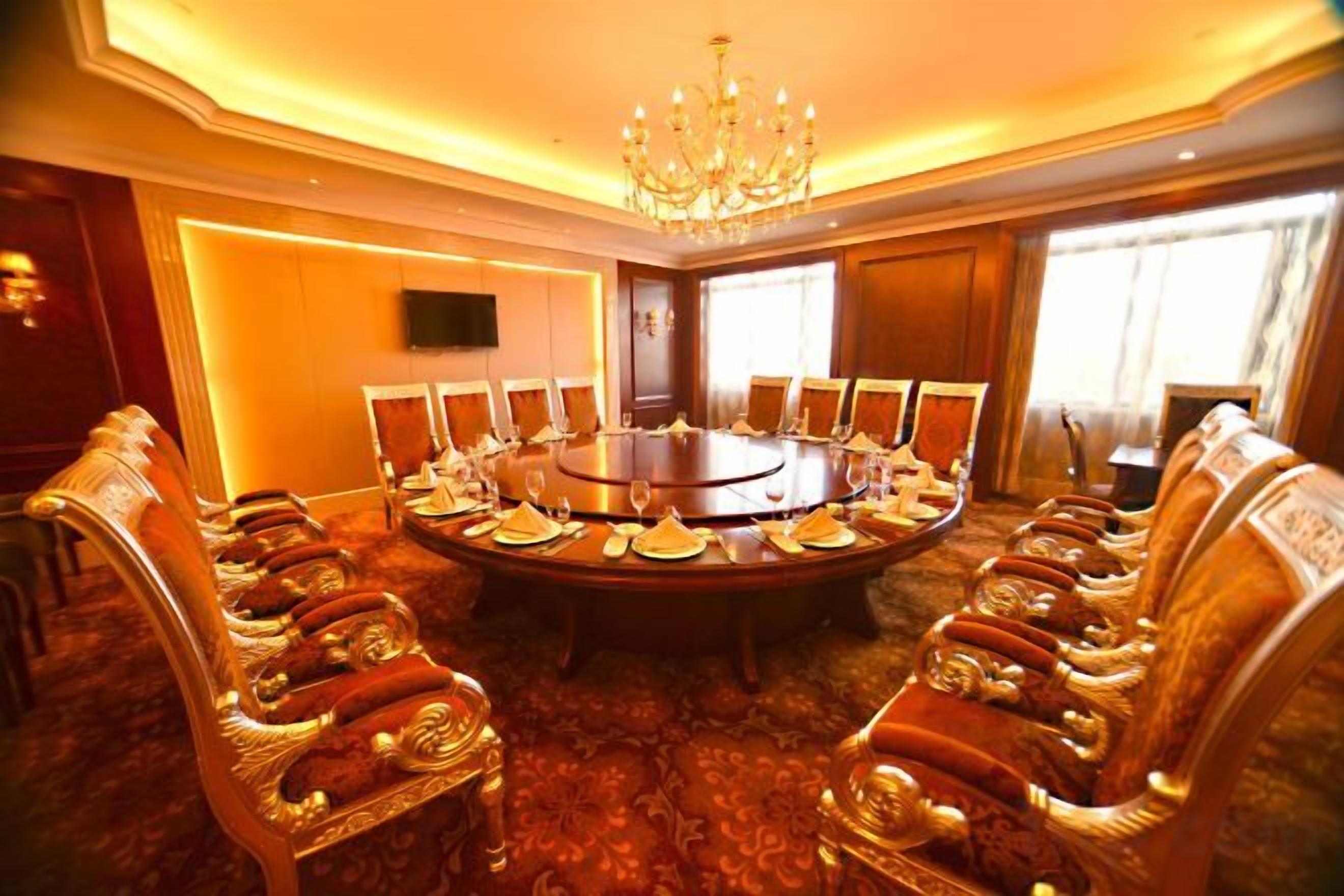 A conference room with a round table, ornate chairs, a chandelier, and a television on the wall.