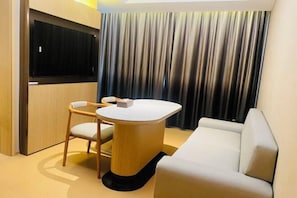 A modern hotel room with a round table, a sofa, a TV mounted on the wall, and a curtain backdrop.