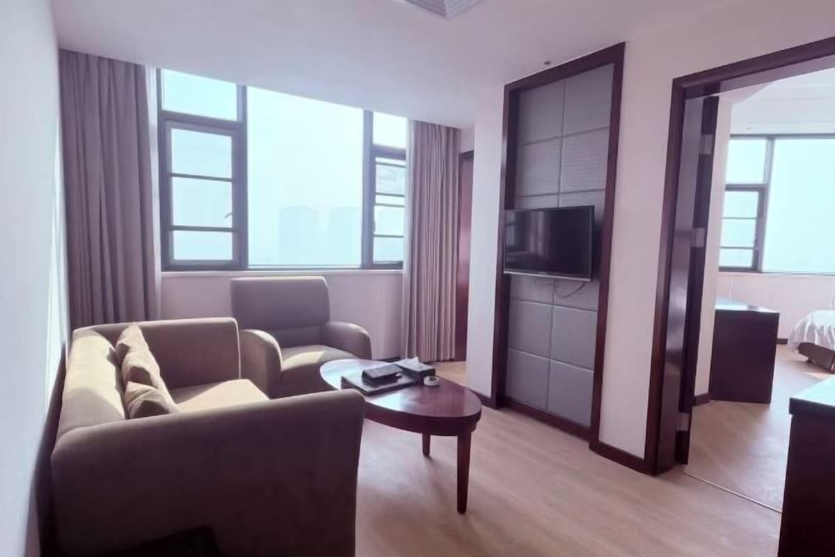 A hotel room with a sofa, armchairs, a small table, a TV, and a sliding door leading to another room.