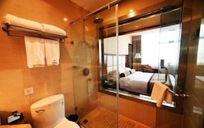 A modern bathroom with a glass shower enclosure, a toilet, and a large mirror reflecting a bedroom with a bed and a nightstand.