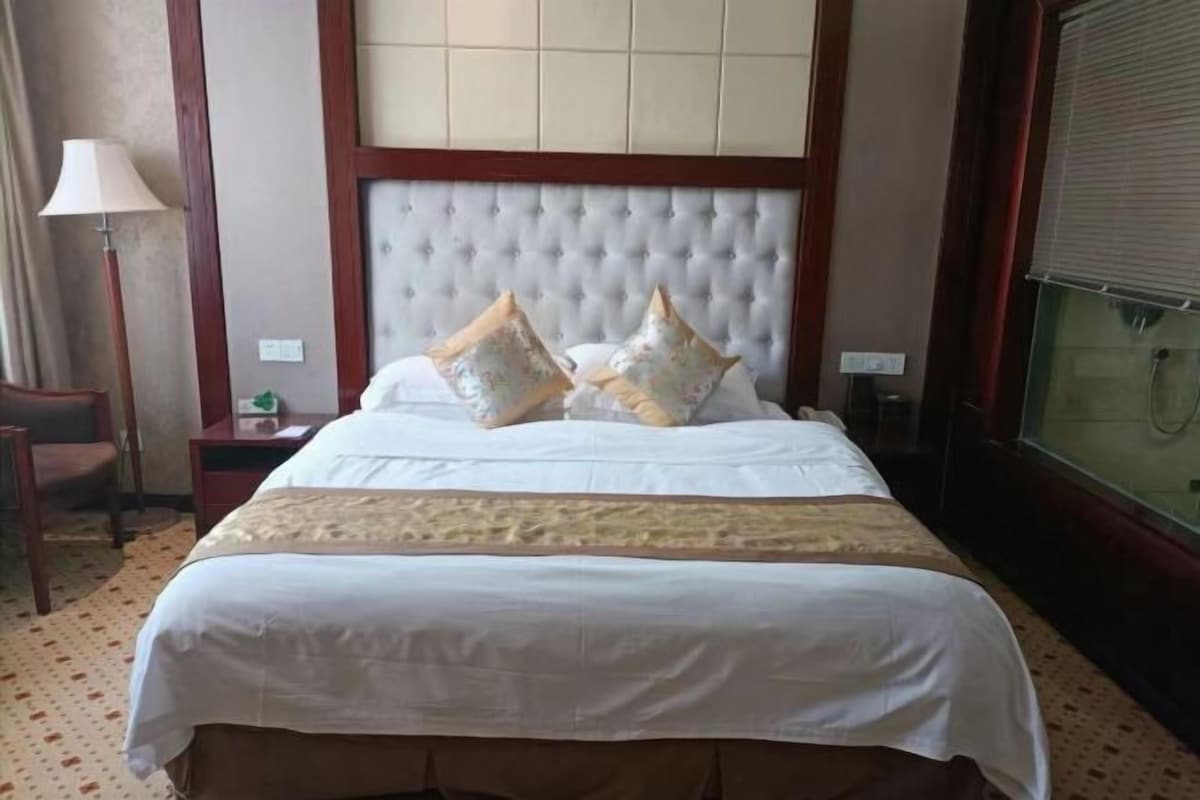 A hotel room with a large bed, a nightstand, a lamp, and a window with blinds.