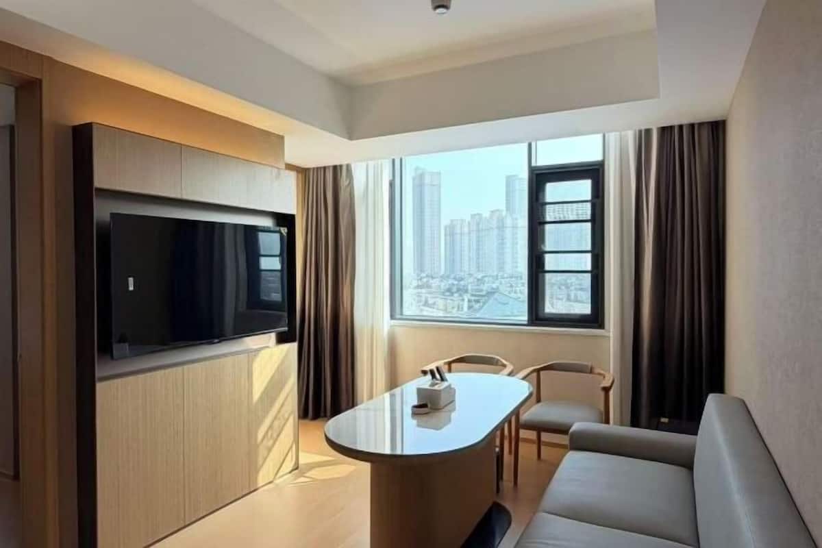 Una habitación de hotel moderna con un televisor de pantalla plana montado en un panel de madera, una pequeña mesa redonda con una silla, un sofá y un amplio ventanal con vistas a la ciudad.