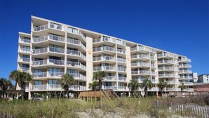 A multi-story hotel with balconies, located near a beach with sandy terrain and sparse vegetation.