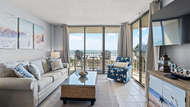 A spacious living room with a sofa, a coffee table, and a blue chair with star patterns. A large window offers a view of the ocean.