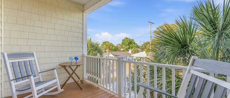 A wooden porch with rocking chairs and a small table, overlooking a garden with palm trees.
