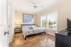 Un dormitorio con cama, mesita de noche, lámpara, ventilador de techo, ventana con persianas y un mueble para televisor.