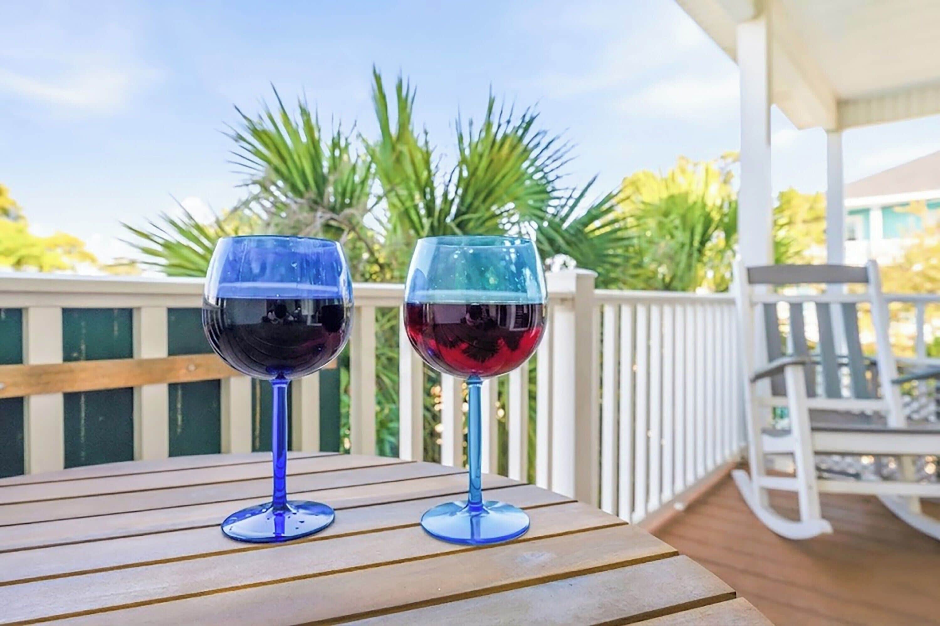 Two wine glasses on a wooden deck with a white railing and greenery in the background.