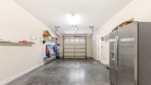 A garage with a large stainless steel refrigerator, a garage door, and various tools and equipment on shelves.