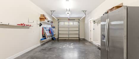 A garage with a large stainless steel refrigerator, a garage door, and various tools and equipment on shelves.