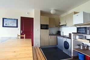 Kompakti keittiö, jossa on mikroaaltouuni, uuni ja pesukone. Keittiössä on puinen ruokapöytä ja punainen ovi.