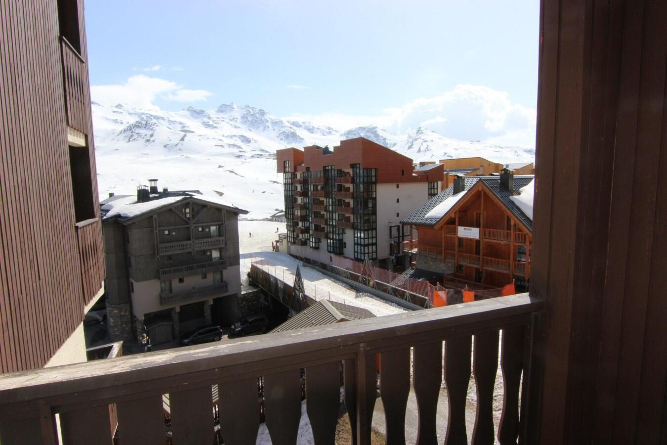 A mountain resort with buildings, including a wooden lodge and a modern multi-story building, with snow-covered mountains in the background.