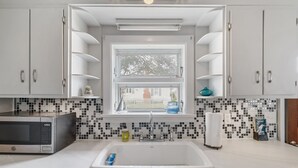 A kitchen with white cabinets, a microwave, a sink, and a window with a view of a house. - Bayside Cottage steps from Rock Hall Harbor (Rock Hall)