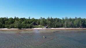 En strand med klart vand, et skovområde og en bygning i baggrunden.