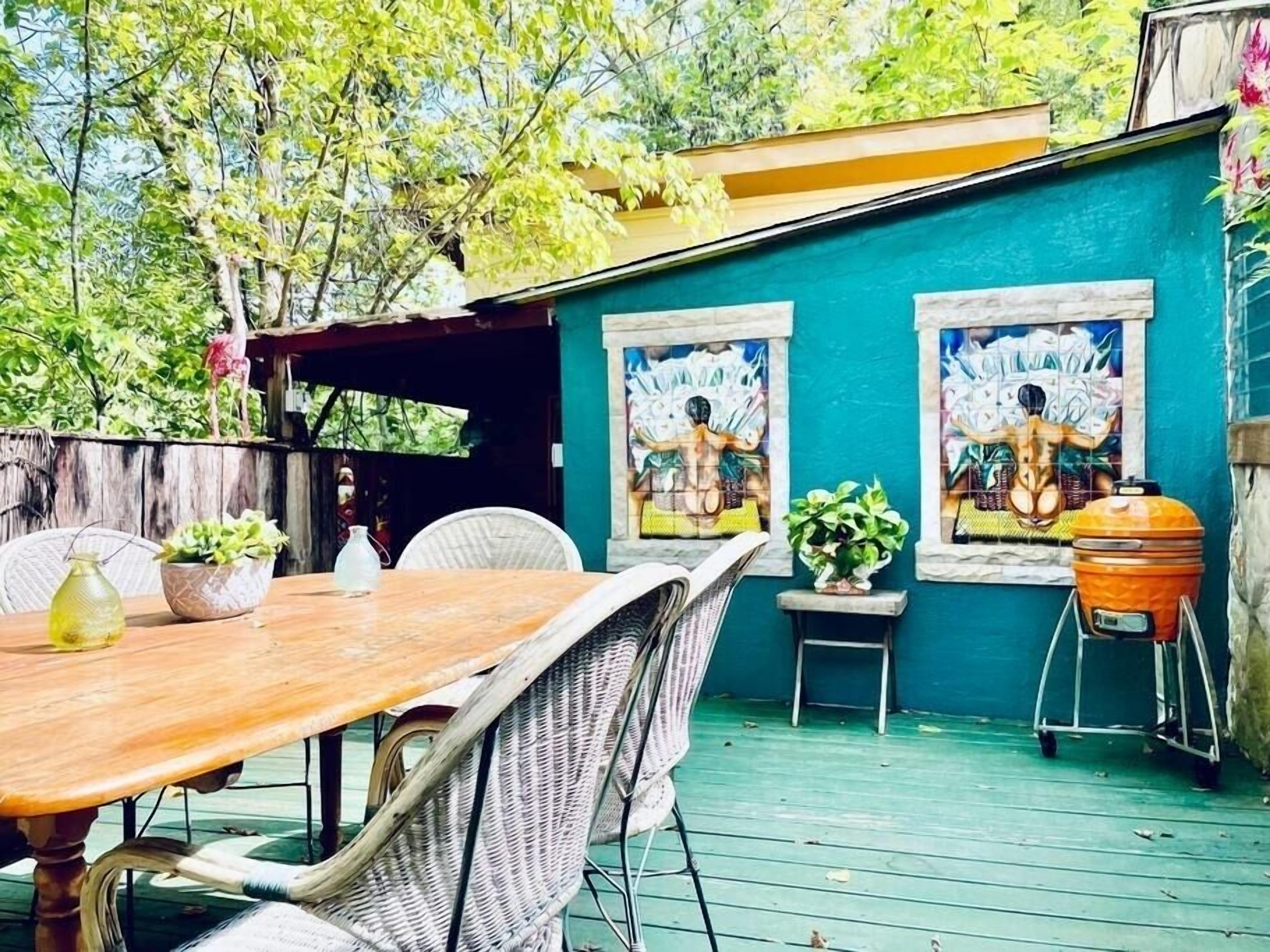 Een terras buiten met een houten tafel en rieten stoelen, een barbecue en twee schilderijen in lijsten aan een muur in teal.
