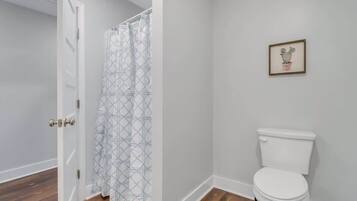 Et badeværelse med et hvidt toilet, et brusebadeforhæng med et geometrisk mønster og et billede i ramme på væggen.