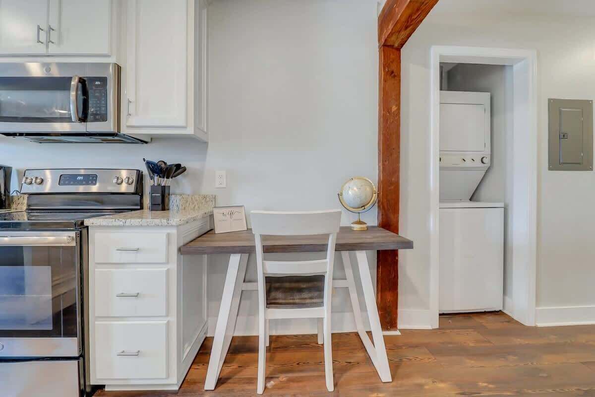 Ett kök med vita skåp, en mikrovågsugn, en spis och ett matbord i trä med en stol.