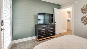 Quarto com cama, penteadeira, espelho e porta que dá para outro cômodo.