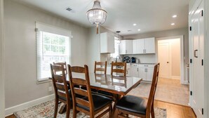 Uma sala de jantar com mesa de vidro e cadeiras de madeira, um candelabro e uma cozinha ao fundo.