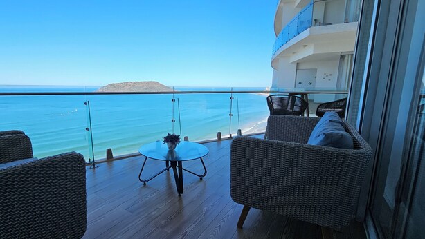 Un balcon agrémenté de meubles en osier, d’une table en verre et offrant une vue sur la mer et une île lointaine.