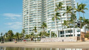Un grand hôtel avec de nombreux balcons, situé près d’une plage bordée de palmiers et dotée d’un rivage sablonneux.