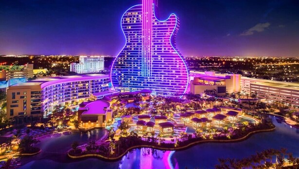 A hotel with a guitar-shaped building illuminated in neon lights, surrounded by a landscaped area with pools and a marina.
