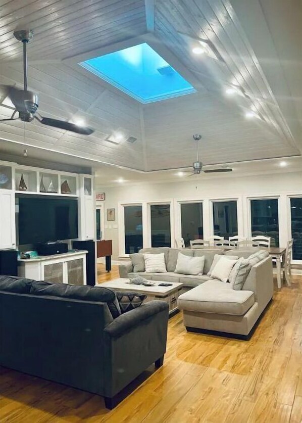 Un salon spacieux avec un plafond haut, une lucarne, un grand canapé, une table basse et un coin repas comprenant une table et des chaises.
