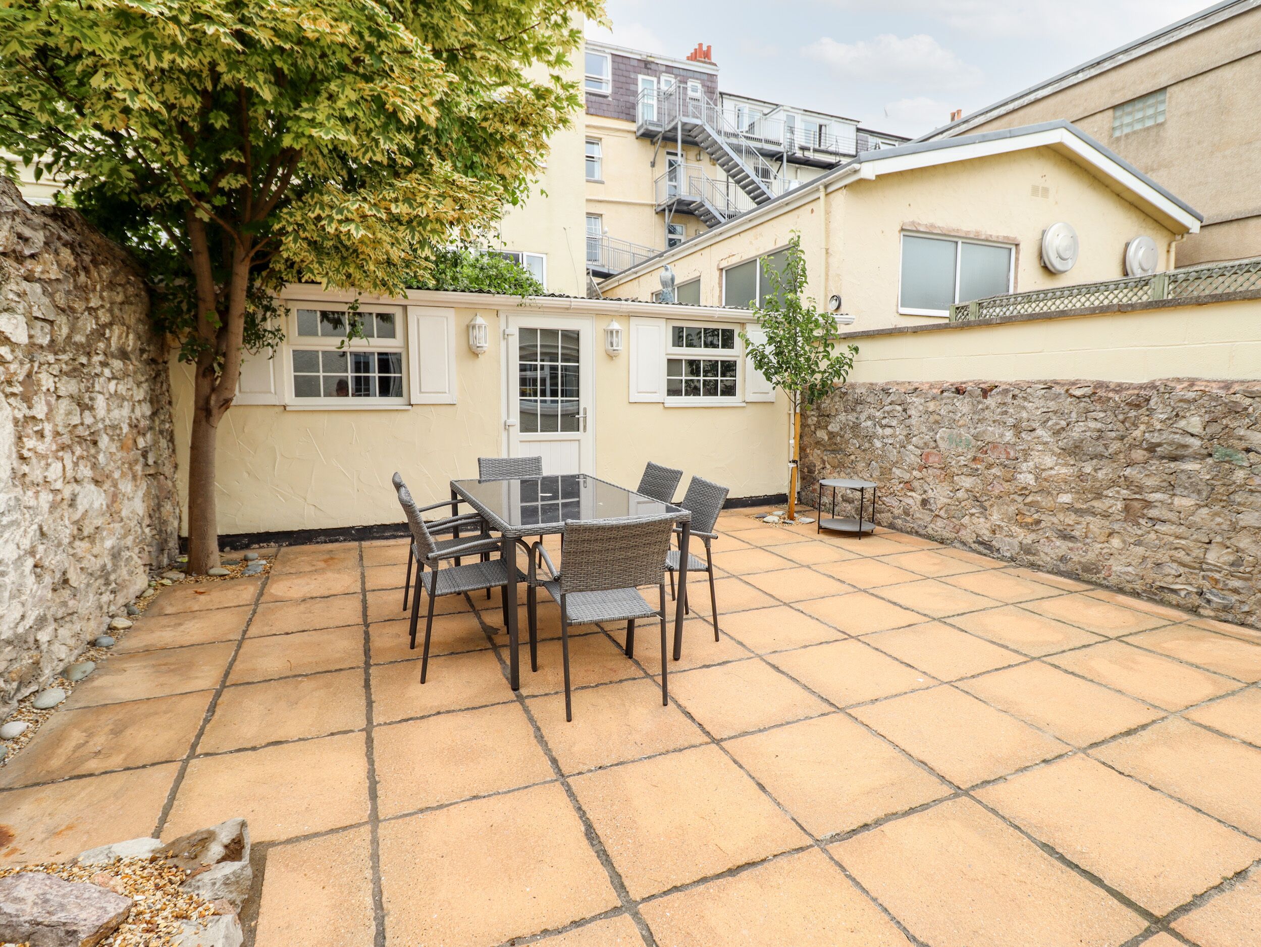 A patio with a table and chairs, a stone wall, and a building with a window and a door in the background.
