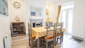 Un comedor con una mesa preparada para cuatro personas, rodeada de sillas. A la izquierda hay una estantería y, al fondo, una chimenea.
