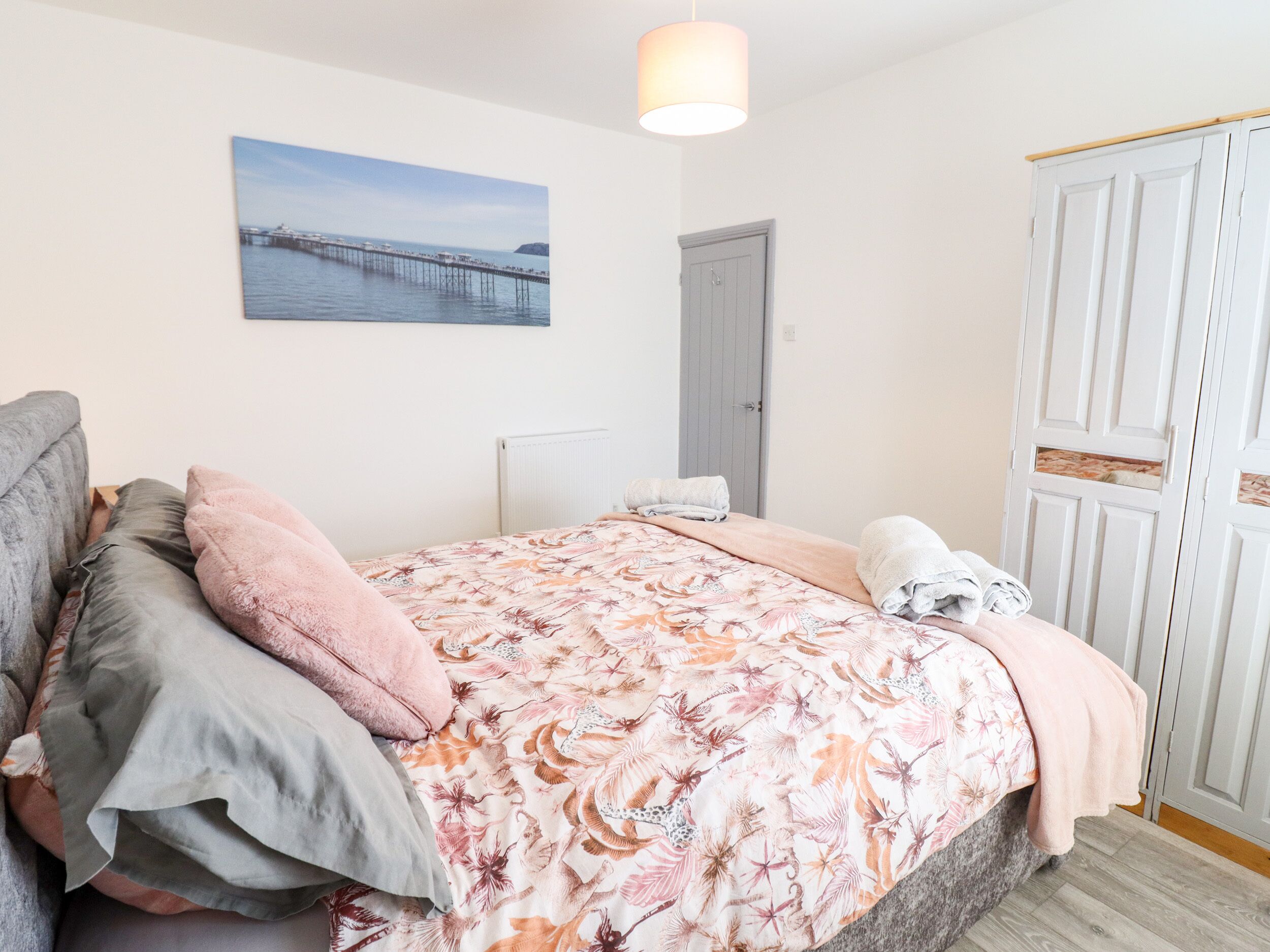 Une chambre à coucher avec un lit, des oreillers, un tableau représentant une jetée, une armoire et un radiateur.