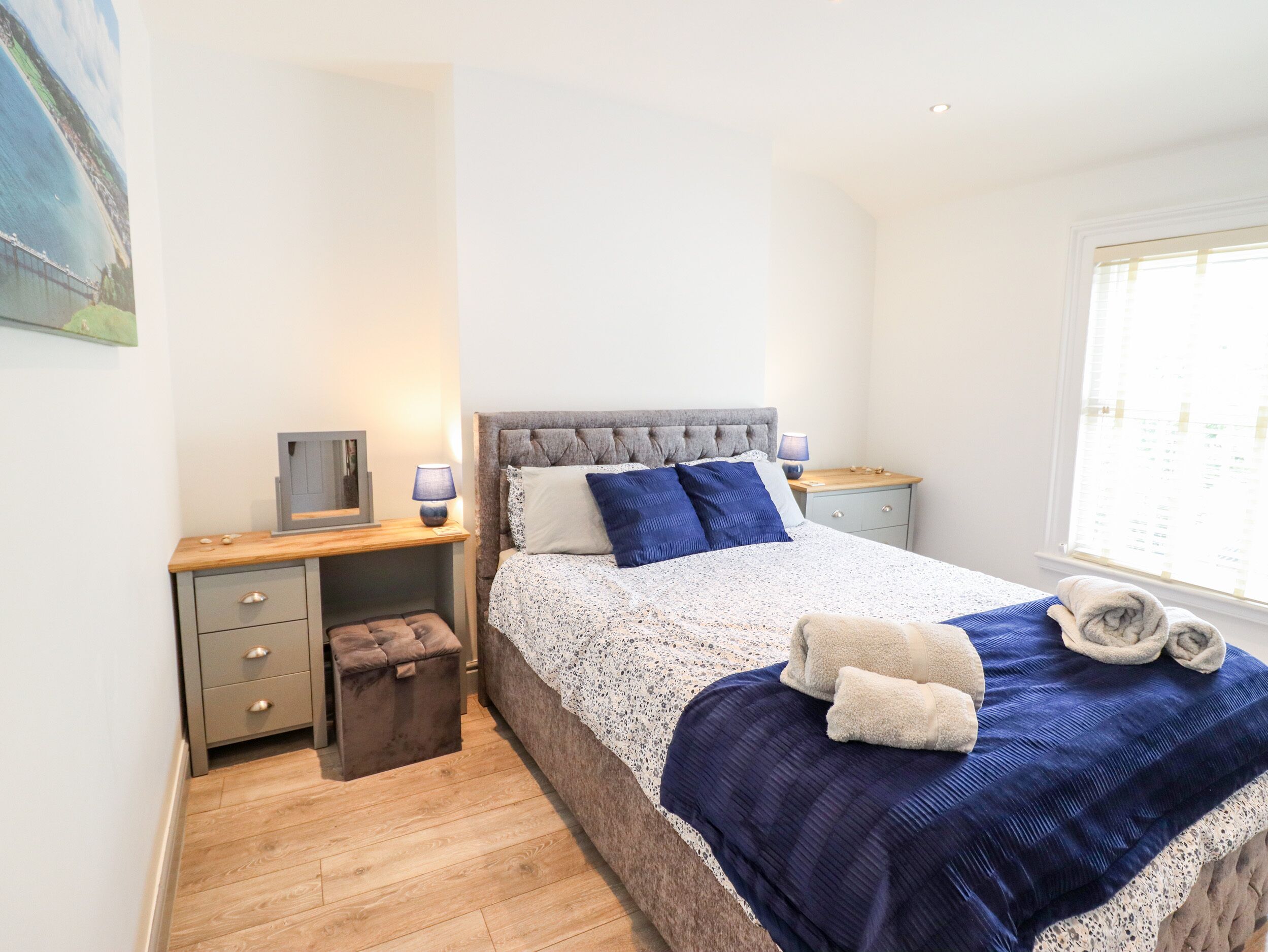 A bedroom with a bed, bedside tables, a small stool, a window, and a painting on the wall.