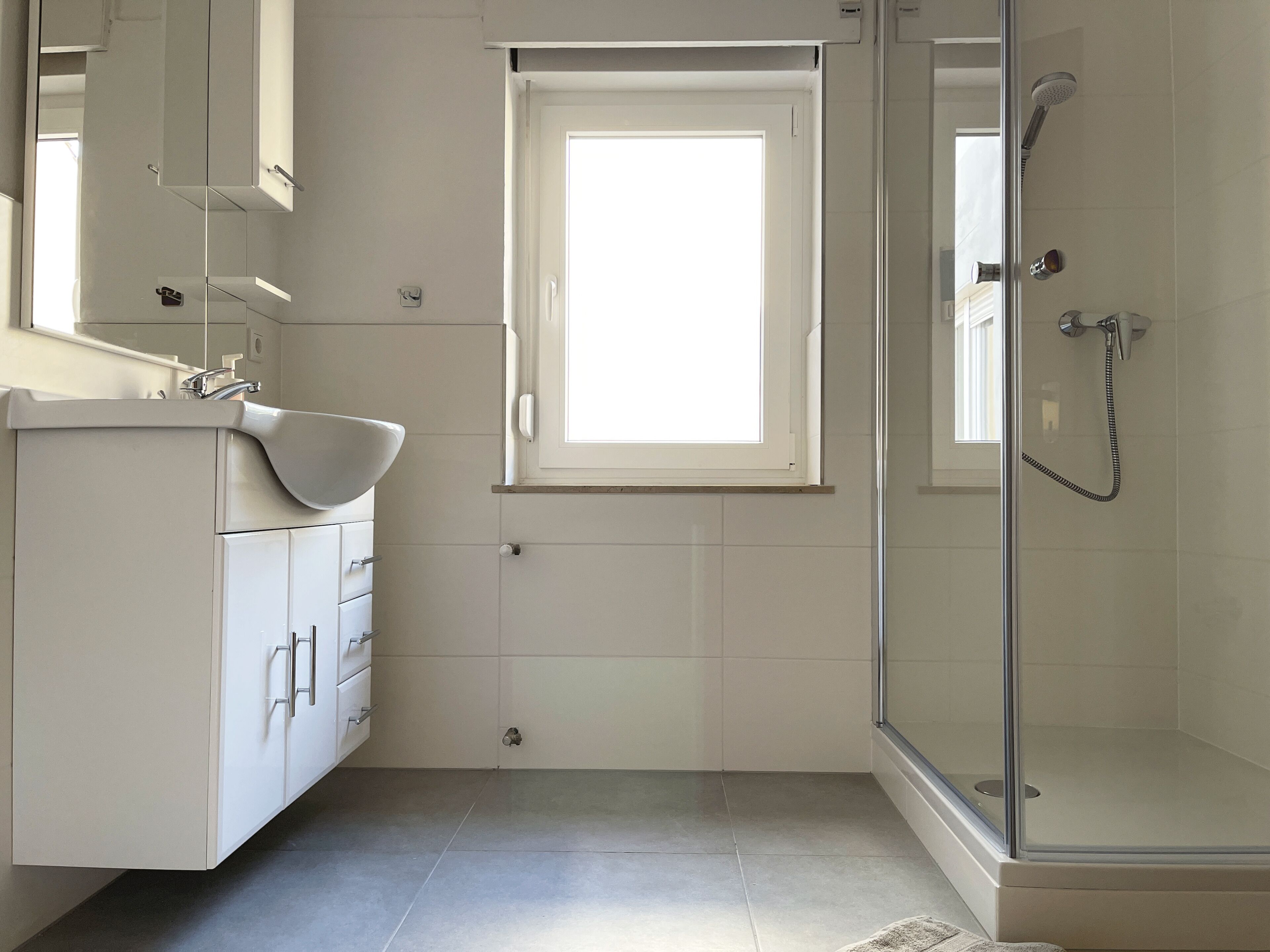 Een moderne badkamer met een douche met glazen wand, een wit wastafelmeubel en een raam voor natuurlijk licht.