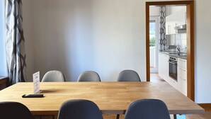 A modern dining area with a wooden table and grey upholstered chairs. A sign is visible on the table, and a kitchen is seen through an open doorway.