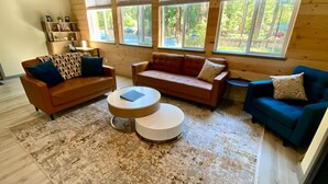 A cozy living room with two sofas, a coffee table, and a bookshelf. Large windows provide a view of the outdoors.