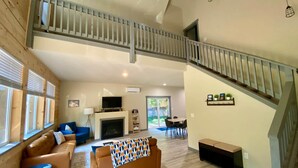 A spacious living area with a high ceiling, a fireplace, a staircase, and a dining area.