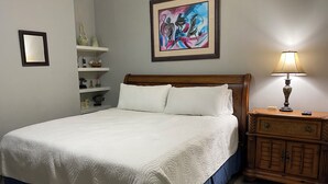 A neatly made bed with a white comforter and pillows, a wooden nightstand with a lamp, a framed abstract painting on the wall, and a shelf with decorative items. - 2 BR/ 2BA Condo1 King /2 Full beds Sleeps 6 close to OWA & Foley Tourism Complex (Foley)