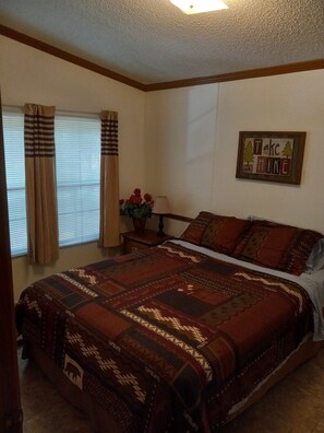 A bedroom with a large bed, a nightstand with a lamp, a window with curtains, and a wall decoration.