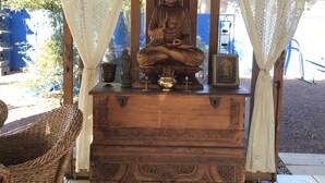 A wooden statue of Buddha on a pedestal, surrounded by white curtains, with a wicker chair and a tiled floor. - The Bali House Experience: Enjoy The Peace (Tucson)