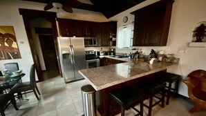 A modern kitchen with dark wooden cabinets, a stainless steel refrigerator, a large island with bar stools, and a ceiling fan. - The Bali House Experience: Enjoy The Peace (Tucson)