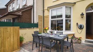 Une terrasse avec une table dressée pour quatre personnes, entourée de chaises, et bordée d’une clôture en bois et de plantes en pot.