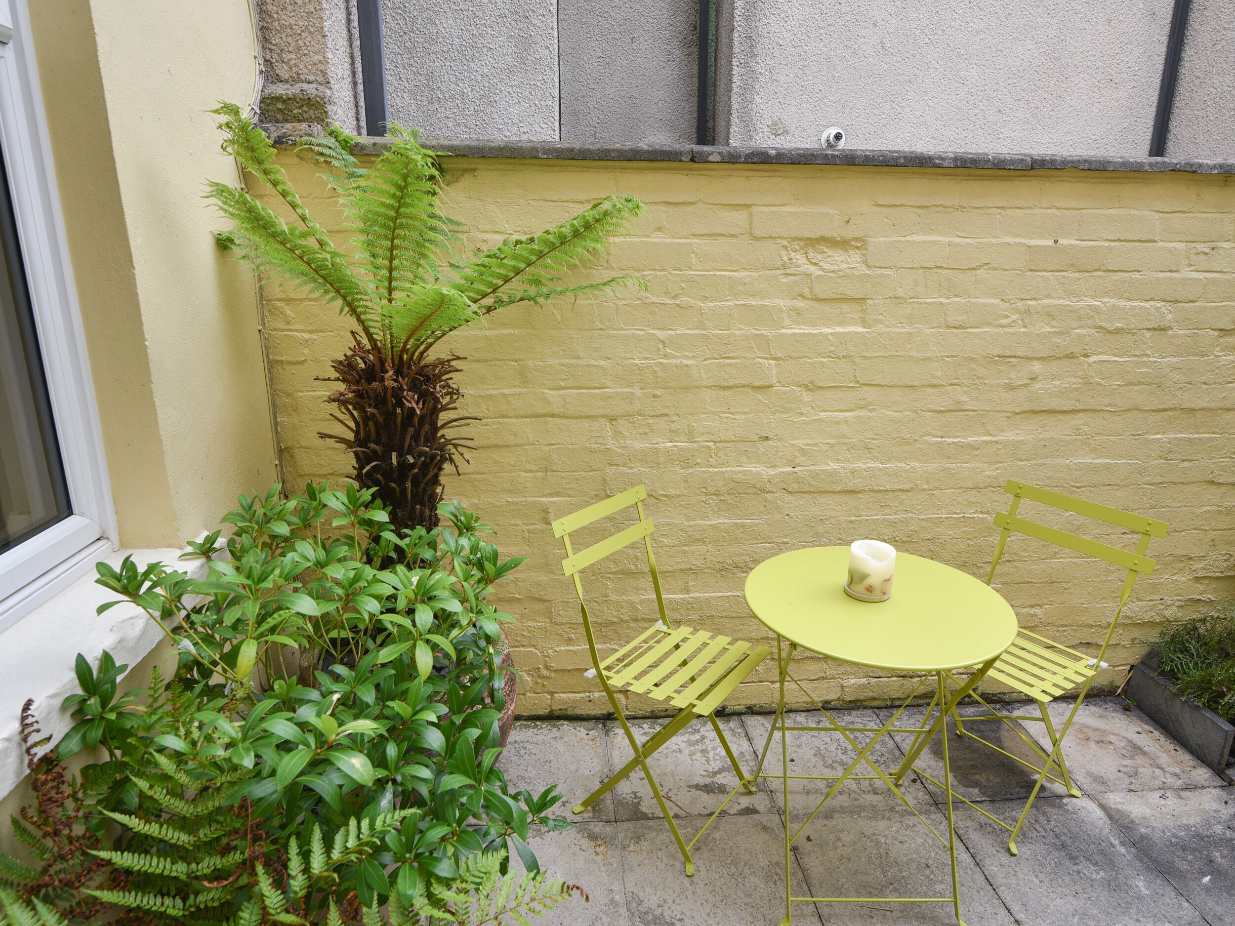A small outdoor seating area with a table and chairs, surrounded by plants and a yellow wall.